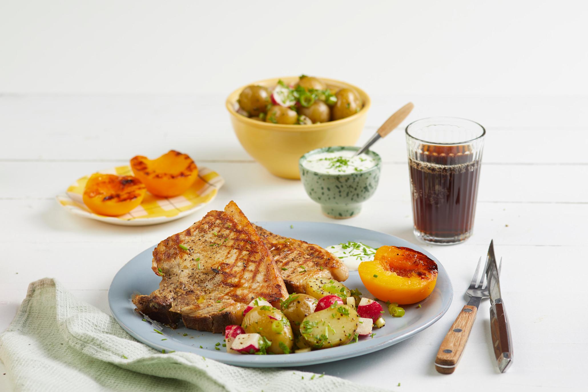 Sommerkoteletter med frisk potetsalat og grillet fersken, servert på lyseblå tallerken med litt hvitløksdressing. Små skåler og fat i bakgrunnen med hvitløksdressing, potetsalat og grillet fersken.