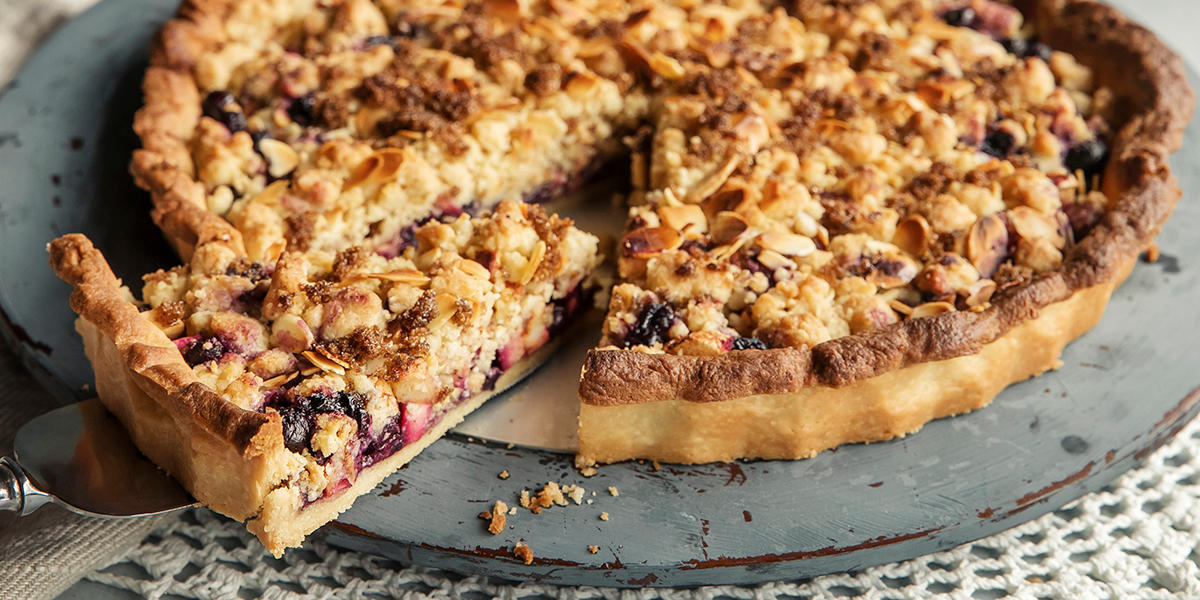 Høstens beste eplepai med blåbær og smuldretopping, servert på mørkegrått trefat på heklet hvit duk. Et stykke pai er skåret ut, og løftes ut med en kakespade.