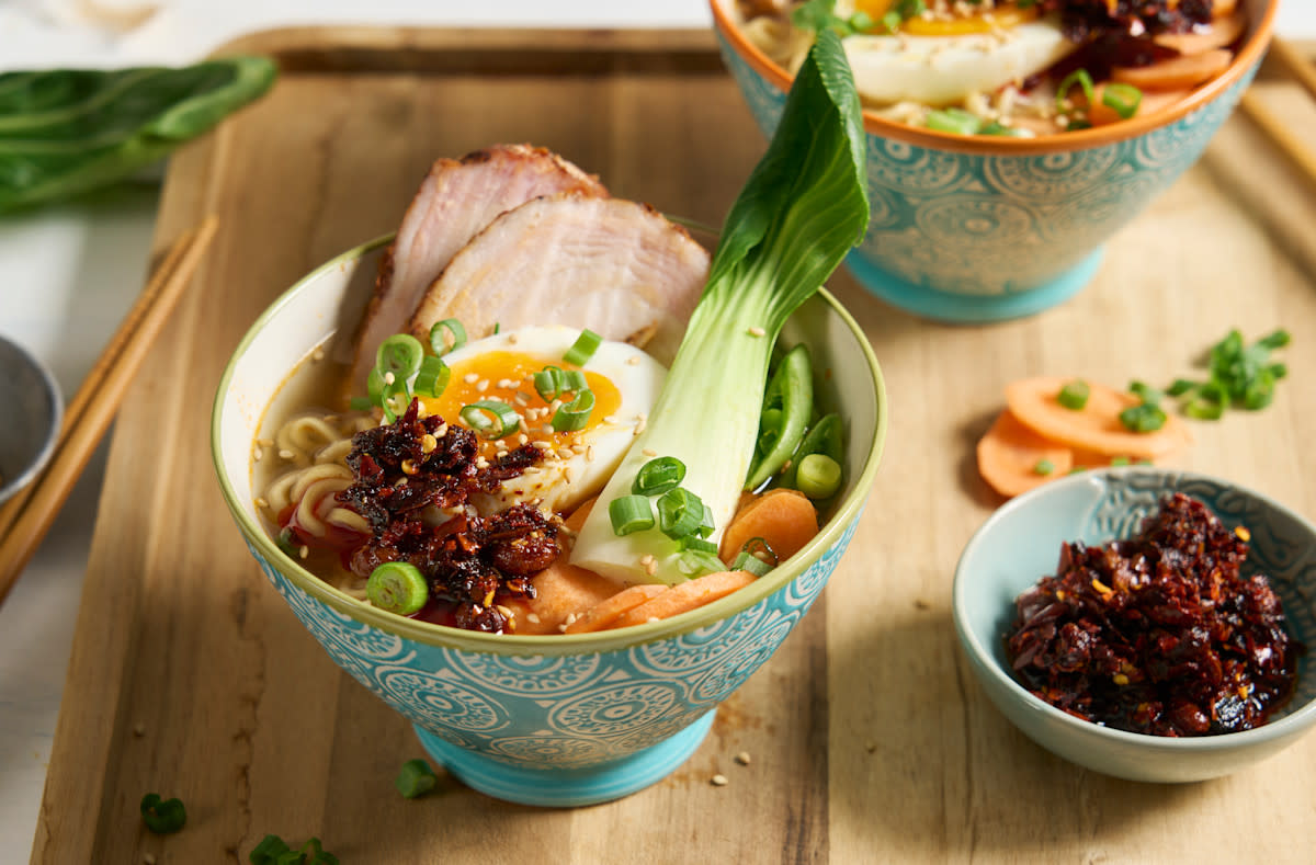 Pork belly ramen med bløtkokt egg og chili crisp servert i turkis skål med gulrot, pak choi, sesamfrø og vårløk. Skålen står på trefjøl med en annen skål med ramen. Liten skål med chilicrisp ved siden av.