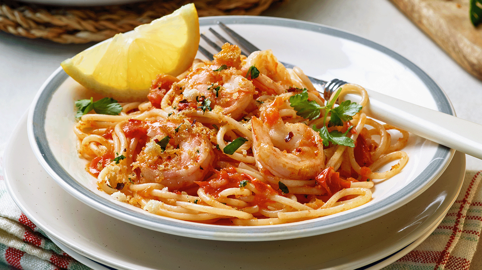 Tallerken med spaghetti og scampi i tomatbasert saus, toppet med fersk persille og brødsmuler, servert med en sitronbåt og hvitt bestikk.