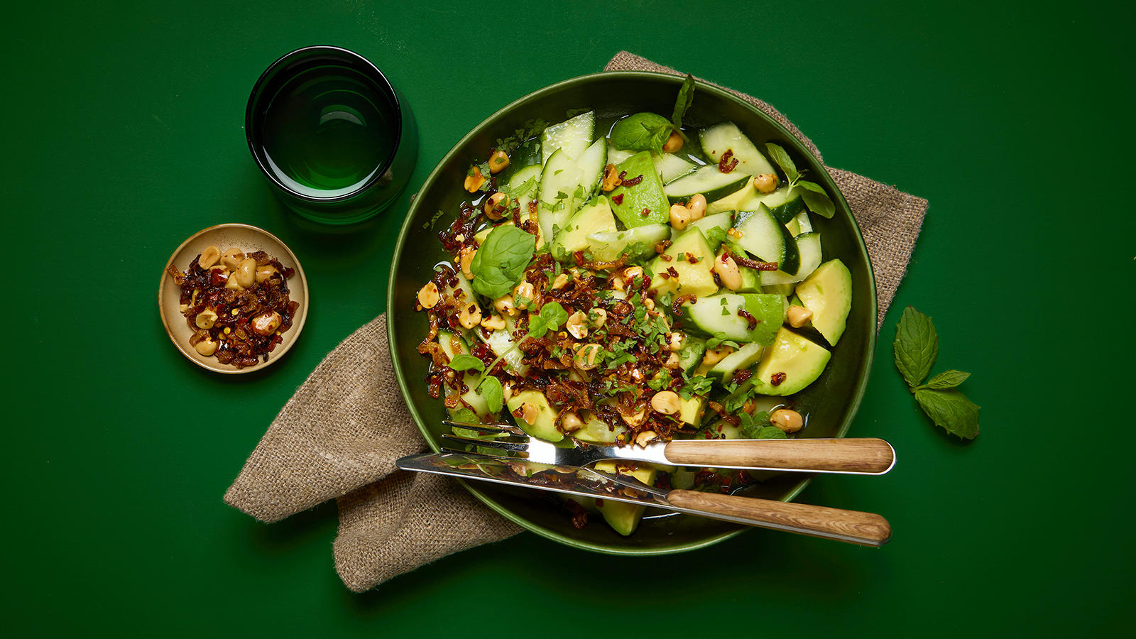 Agurk- og avokadosalat med sprø peanøttopping servert i mørkegrønn bolle trebestikk på siden.