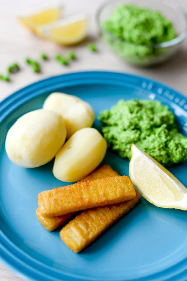 Fiskepinner med hjemmelaget ertepuré, kokt potet og en sitronbåt servert på blå tallerken. Skål med ertepuré i bakgrunnen.