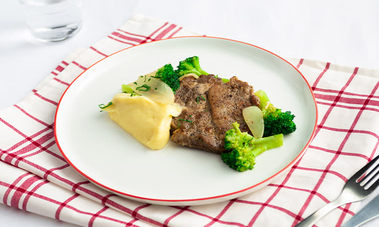 Svinekoteletter med potetmos og brokkoli servert på en hvit tallerken med rød kant, ligger på et rødt og hvitt rutete håndkle på hvitt middagsbord.