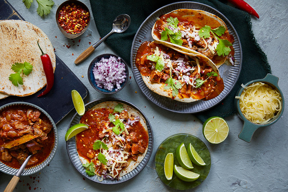 Birria taco med langtidskokt lam servert i tacolefser fordelt utover diverse tallerkner. Små skåler med tilbehør rundt.