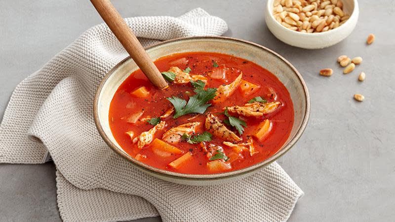 Rød kyllingsuppe med biter av kylling og søtpotet, toppet med ferske urter, i lys suppeskål. Pinjekjerner til pynt ved siden av.