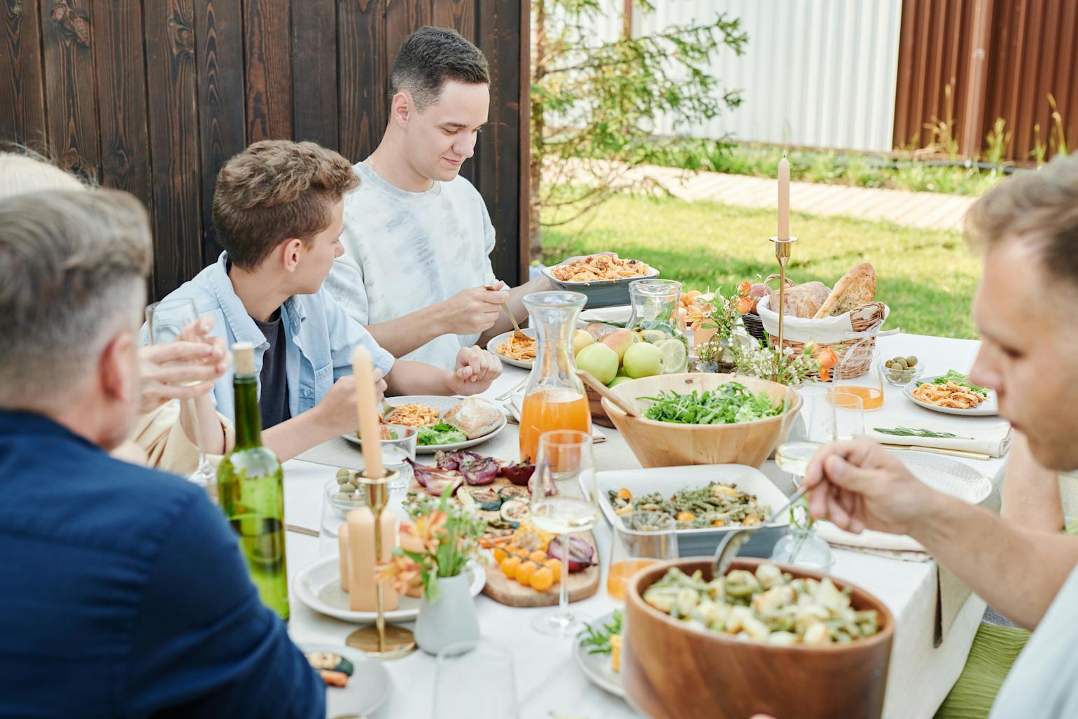 Folk sitter ute rundt et dekket bord, spiser grillmat, salat og brød.