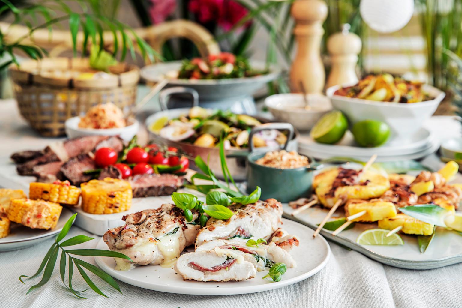 Middagsretter laget på grillen; fylt kylling med spekeskinke og ost, grillet mais, flanksteak med tomater, grillet potetsalat med asparges og grillspyd med kylling, bacon og ananas. Rettene er servert på dekket middagsbord med sommerfølelse.