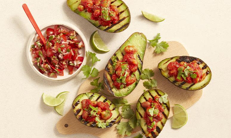 Grillet avokado delt i to, fylt med tomatiserte bønner, ligger på lys trefjøl. Limebåter, fersk koriander og tomatsalsa ved siden av.