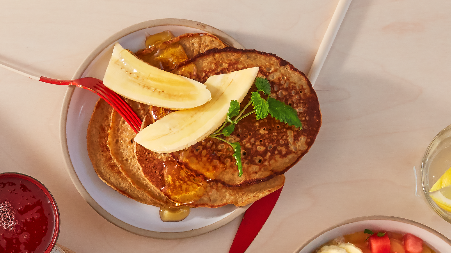 Bananpannekaker med havregryn, servert med fersk banan og lønnesirup på liten hvit tallerken. Lys bakgrunn med sommerlig lys.