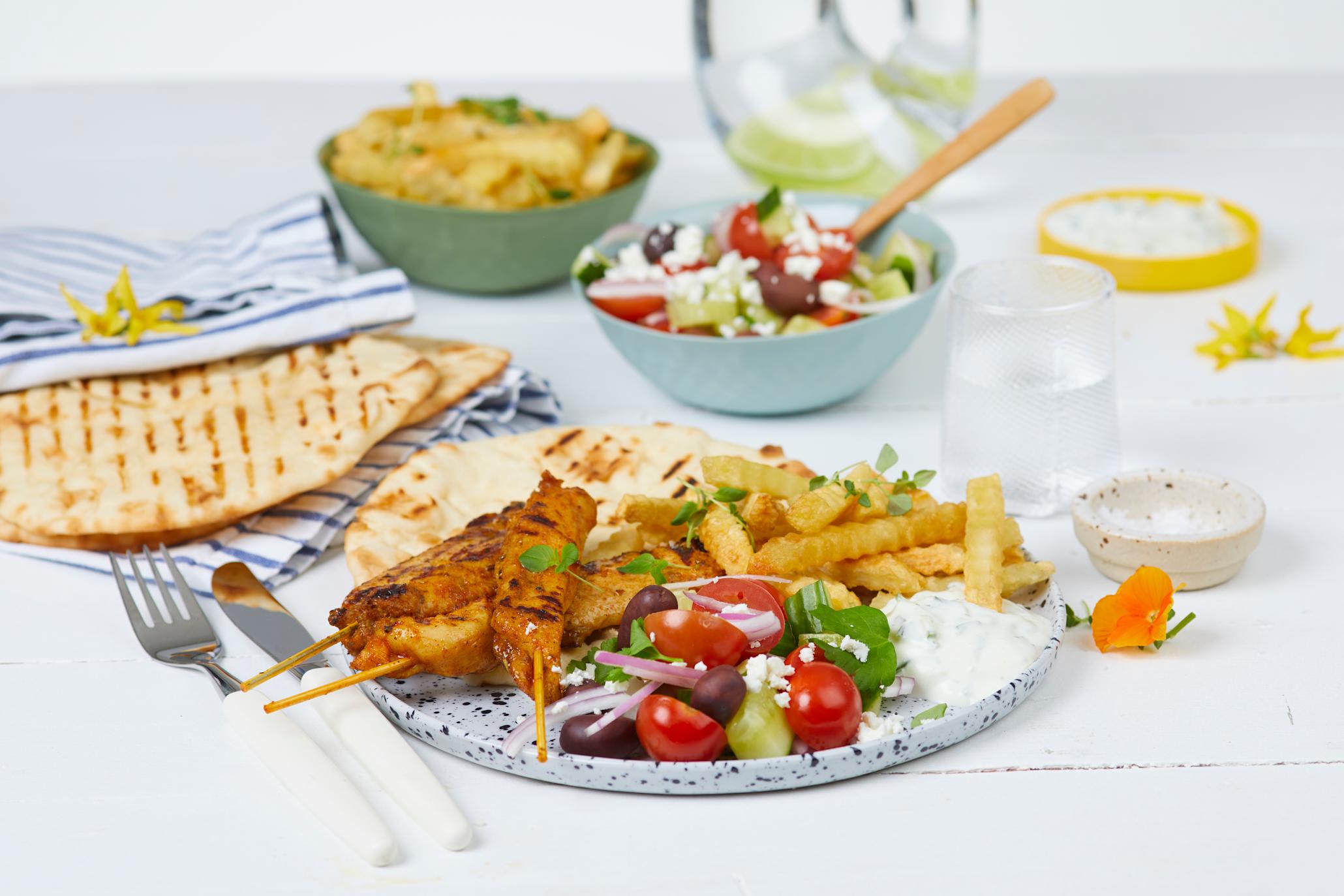 Kyllingspyd Gyros med gresk salat, tzatziki, Coop fries og pita på en lys tallerken med blå splatter foran en skål med salat, fries og fler pitabrød.