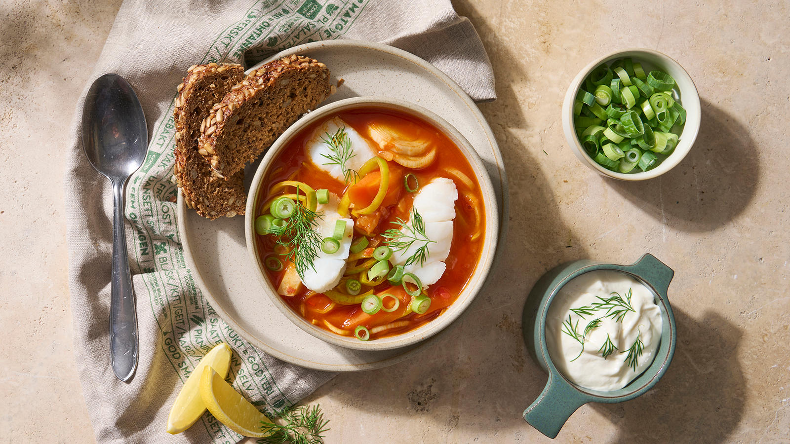 Rød fiskesuppe med torsk og grønnsaker, pyntet med vårløk og dill, og servert i lys skål med grøvbrød på siden.