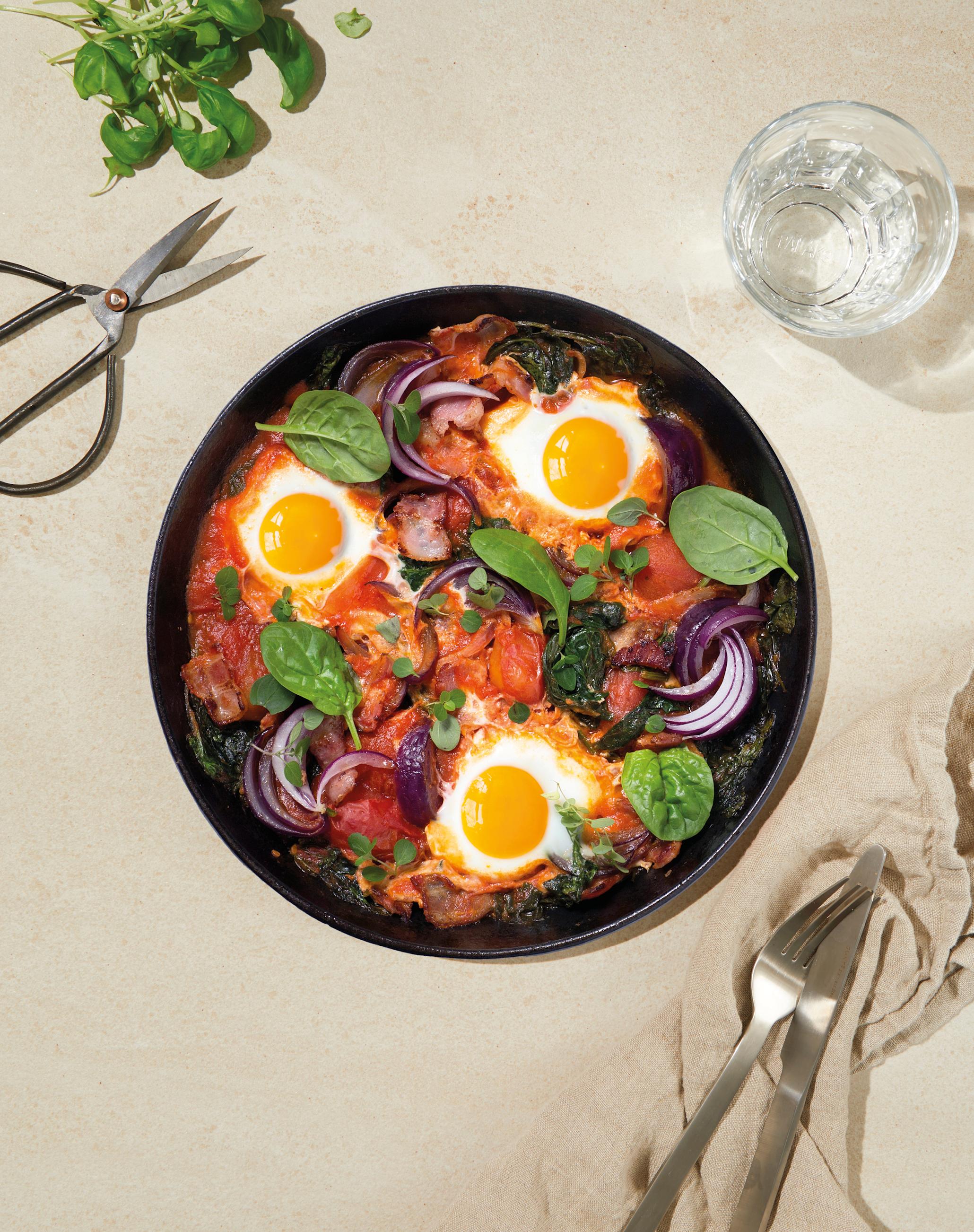 Bakt egg med spinat, tomat, løk og urter servert i ildfast form på beige bakgrunn med linserviett og ferske urter på siden.