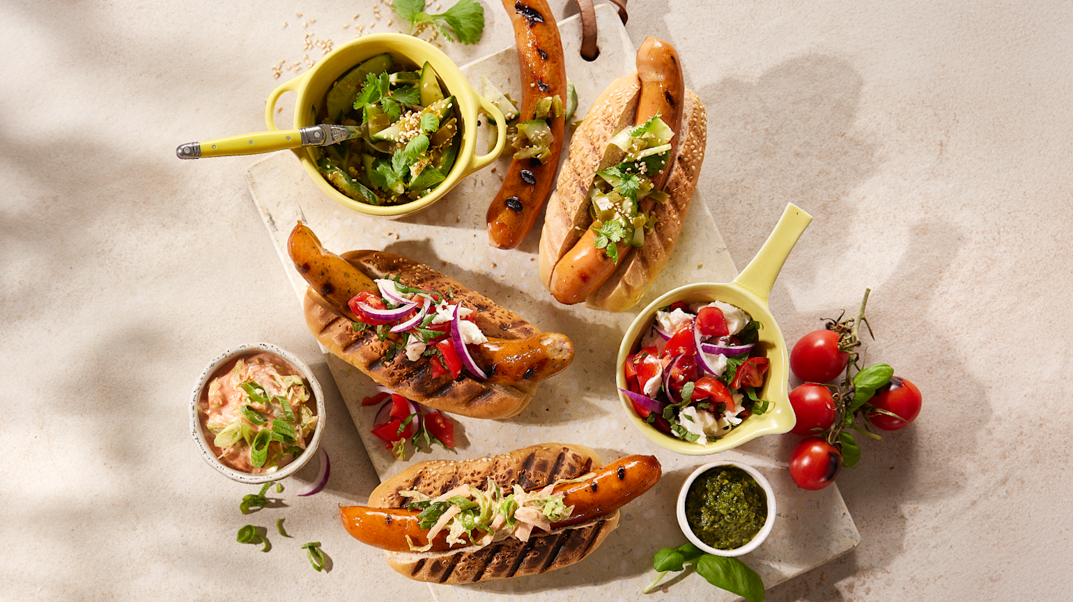 3 forskjellige tilbehør til pølser servert på grillede pølser i brød, og i skåler ved siden av. Pølsene ligger på lys trefjøl med friske urter rundt.