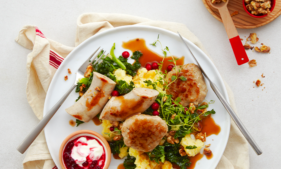 Juletallerken med hjemmelaget mandelpotetmos og hvitløksstekt grønnkål, servert med julepølse, medisterkaker, tyttebærrømme og brun saus på hvit tallerken.