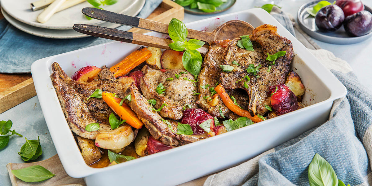 Ovnsbakte nakkekoteletter med småpoteter, friske plommer og gulrøtter servert i hvit firkantet ildfast form med ferske urter strødd over. Formen står på dekket middagsbord, med en skål ferske røde plommer i bakgrunnen.
