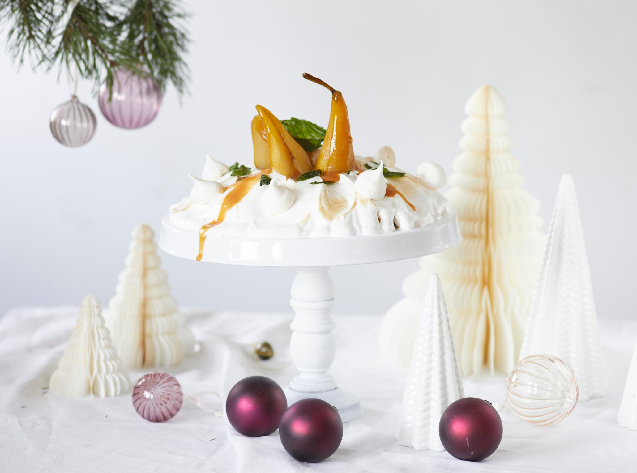 Julepavlova med krydderkokte pærer og hjemmelaget karamellsaus, servert på hvitt stettfat på julepyntet hvitt bord med julekuler og julepynt i gull og lilla.