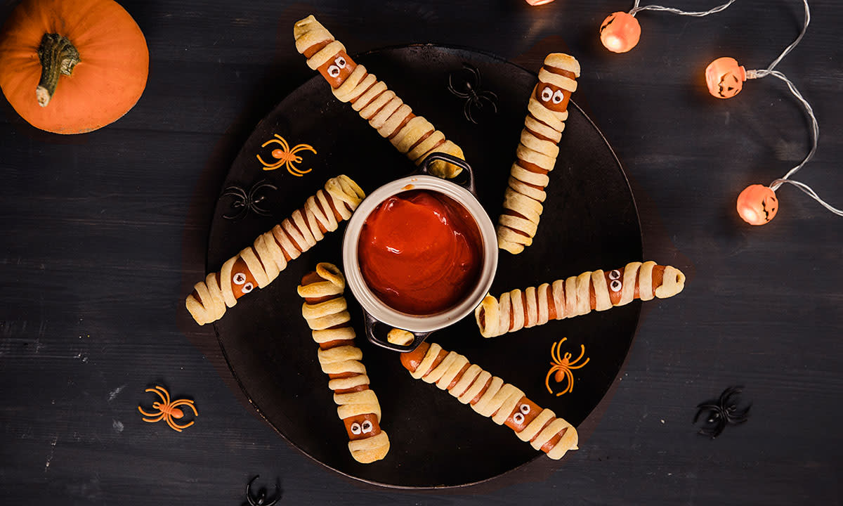 Skumle mumiepølser pakket inn i pizzadeig, med øyne laget av hvit glasur og rosiner. Servert på svart rund tallerken på mørkt bord, med halloweenpynt rundt.