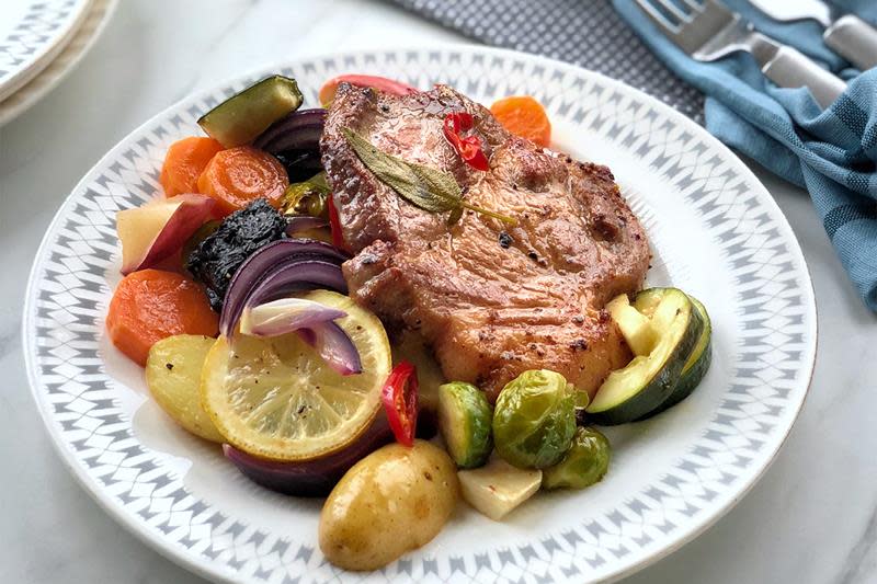 Kotelett servert på en seng av fargerike grønnsaker på hvit og gråstripet tallerken, på dekket middagsbord. Blå serviett ved siden av.