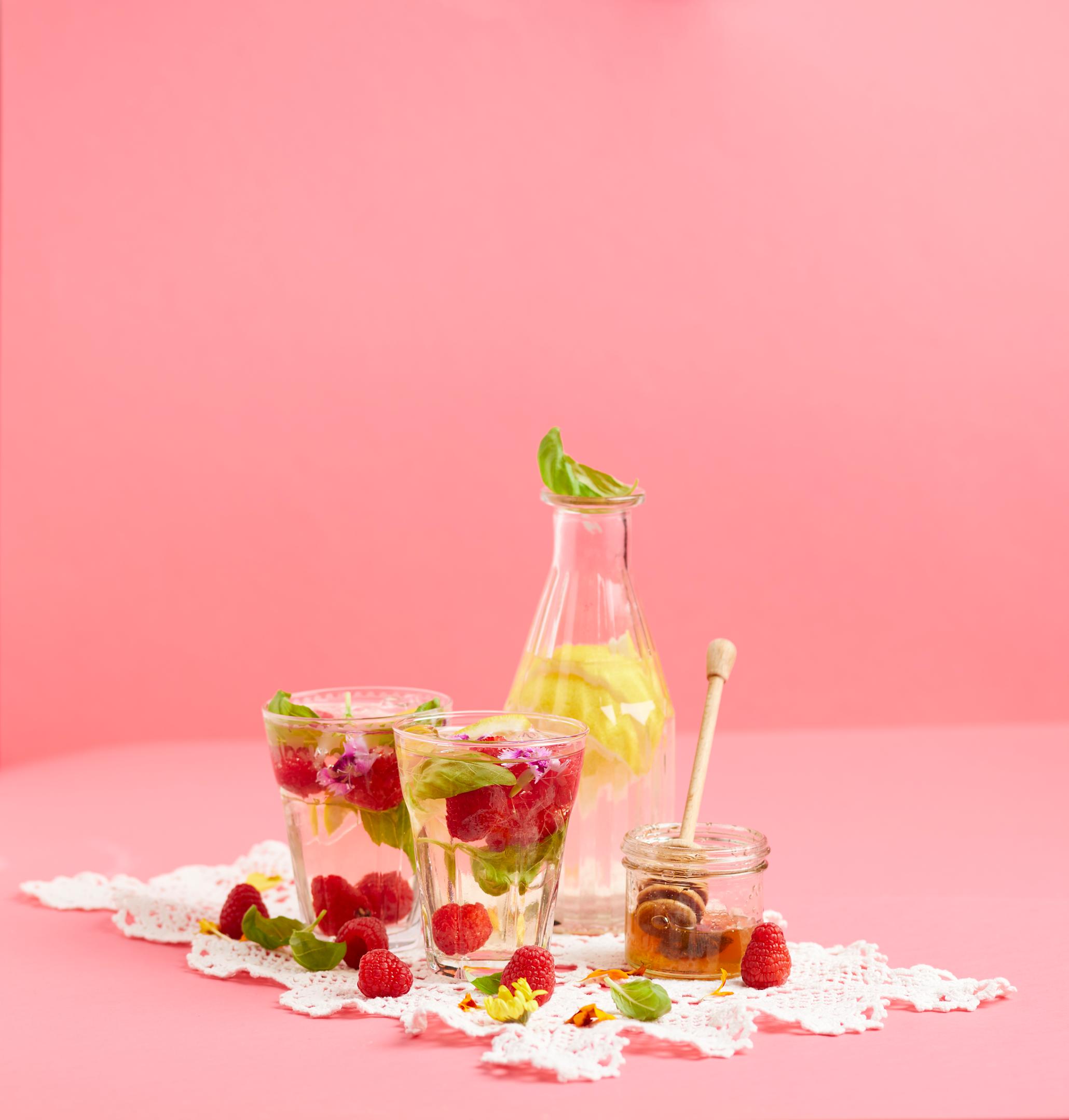 Hjemmelaget iste med kamille, bringebær og sitron servert i glass med friske bringebær, basilikumblader, sitronskiver og lilla blomster. Glassene står på rosa bakgrunn med en karaffel i glass bak, og en krukke honning på siden.