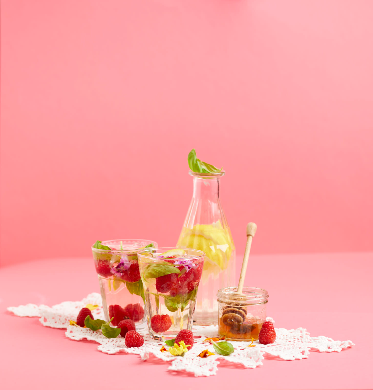 Hjemmelaget iste med kamille, bringebær og sitron servert i glass med friske bringebær, basilikumblader, sitronskiver og lilla blomster. Glassene står på rosa bakgrunn med en karaffel i glass bak, og en krukke honning på siden.