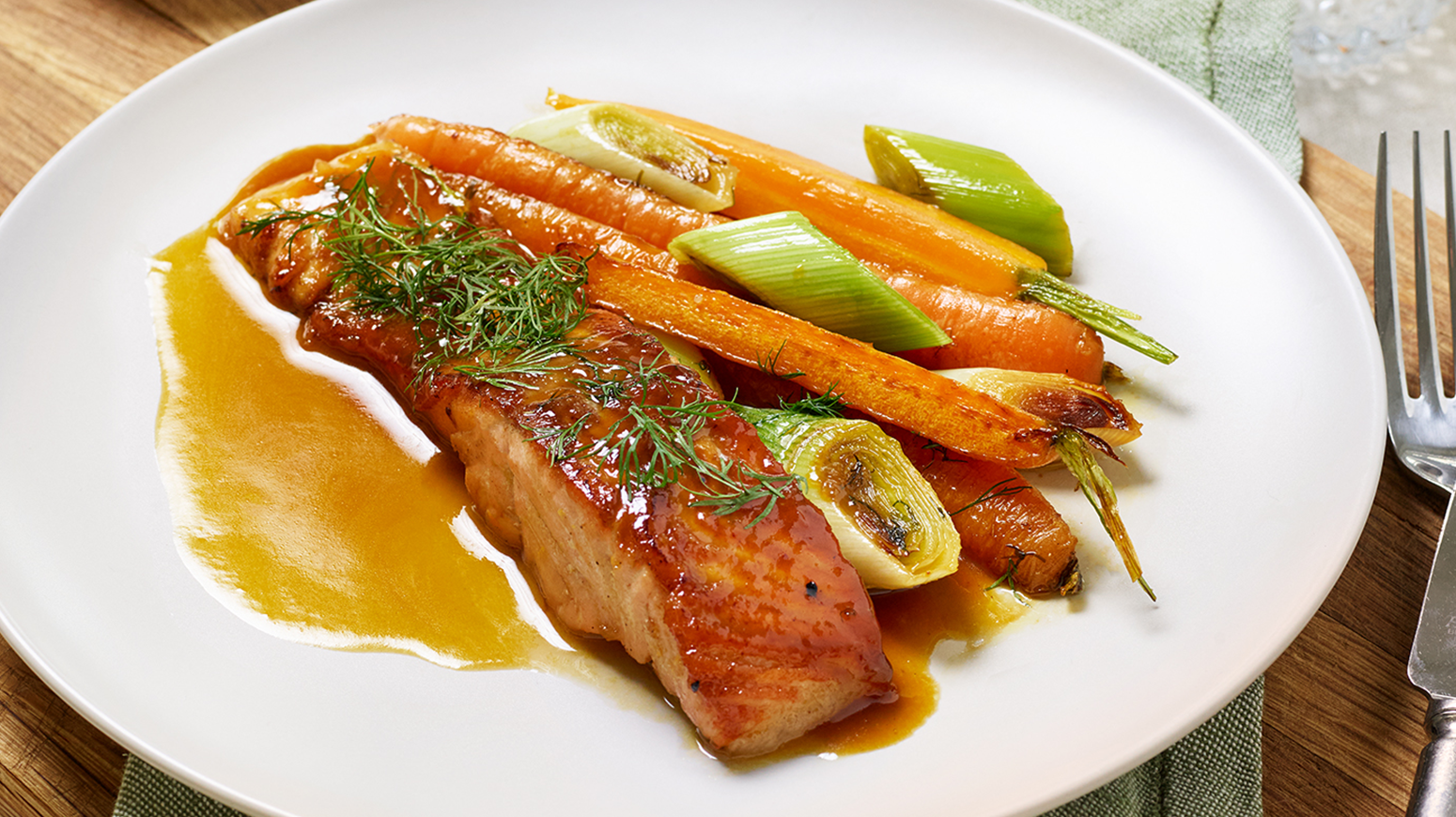 Hvit tallerken med en porsjon Laks toppet med dill og appelsinsaus. Gullrøtter og purre ved siden av.