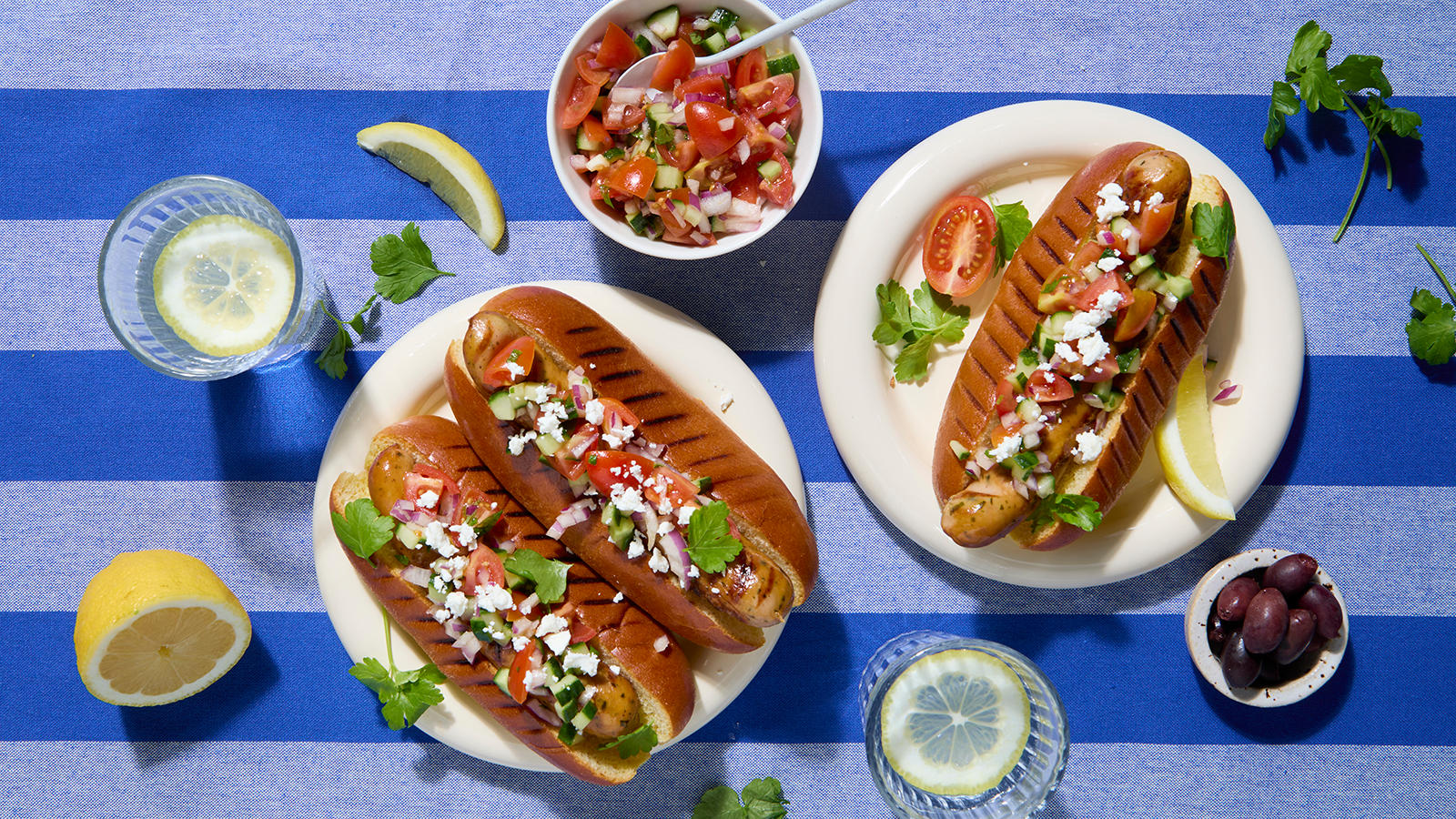 Tasty Garlic pølse med gresk salsa og fetaost, servert i pølsebrød på lyse tallerkner. Skål med gresk salsa, og glass med sitronskiver rundt.