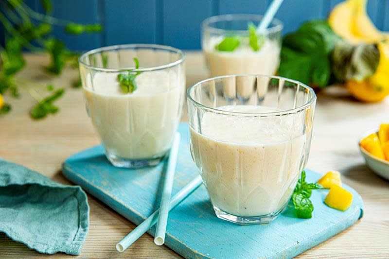 Banansmoothie med mango i to glass står på blå trefjøl med papirsugerør mellom seg. Et glass med smoothie, bananer, mango og ferske urter i bakgrunnen.