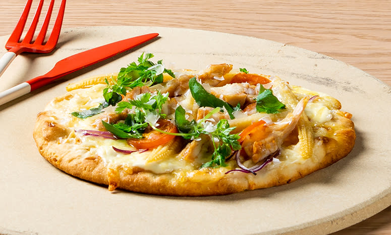 Hvit pizza med kylling, rødløk, tomater og mais servert på en stor skjærefjøl med en rød kniv og gaffel på venstre side