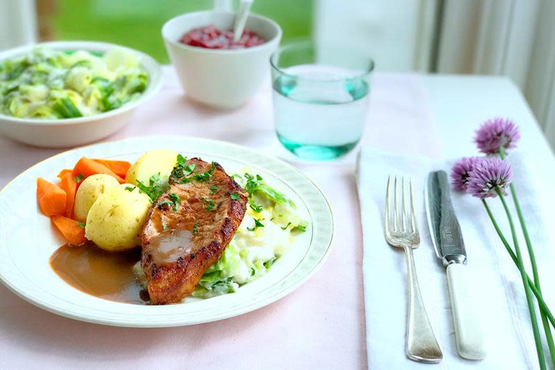 Middagstallerken med røkt svinekam, kålstuing, kokte poteter og gulrot på sommerdekket middagsbord. Skål med kålstuing og skål med tyttebærsyltetøy i bakgrunnen.