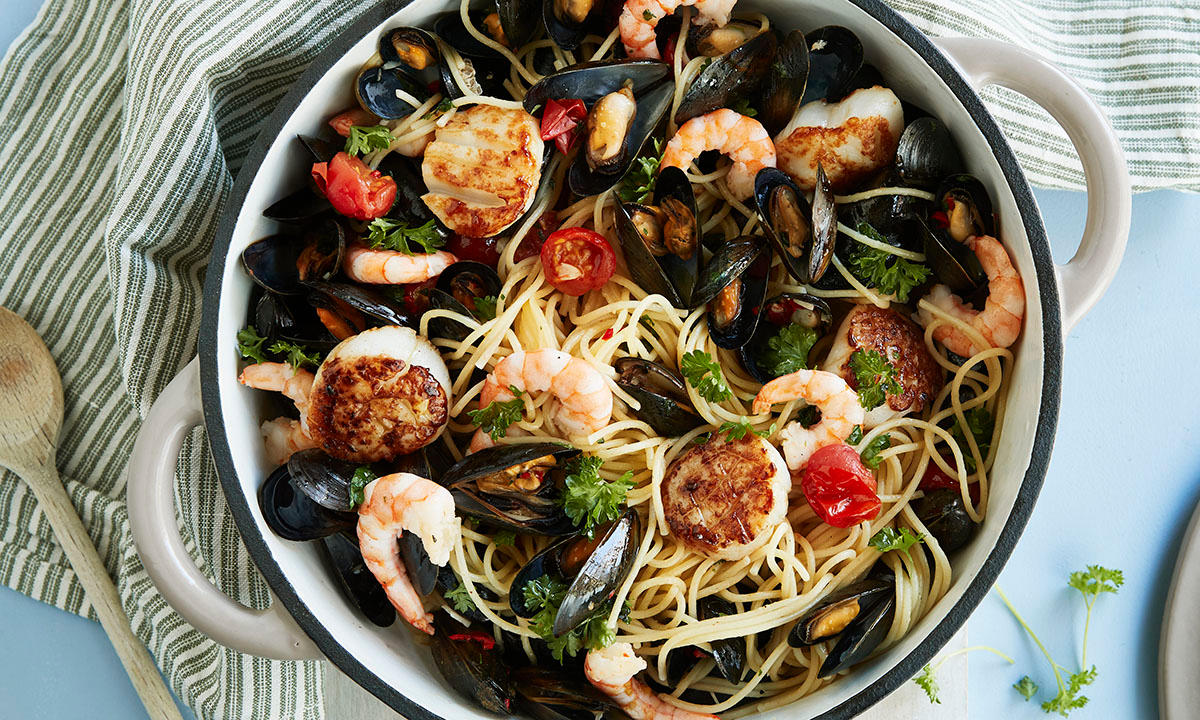 Frutti di mare - italiensk skalldyrpasta servert i hvit kjele med spaghetti kamskjell, blåskjell, reker og ferske urter. Stripete kjøkkenhåndkle og tresleiv på siden.