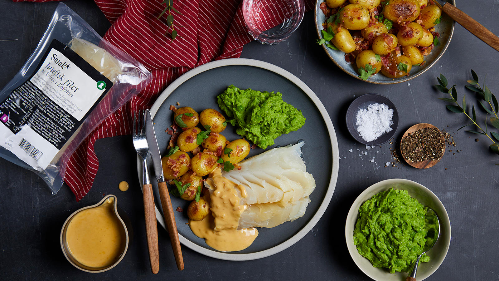 Lutefisk i ovn med baconpoteter, ertestuing og hvitvinssaus servert på svart tallerken med hvit kant. Skåler med baconpoteter og ertestuing, og lite sausenebb med hvitvinssaus på siden.