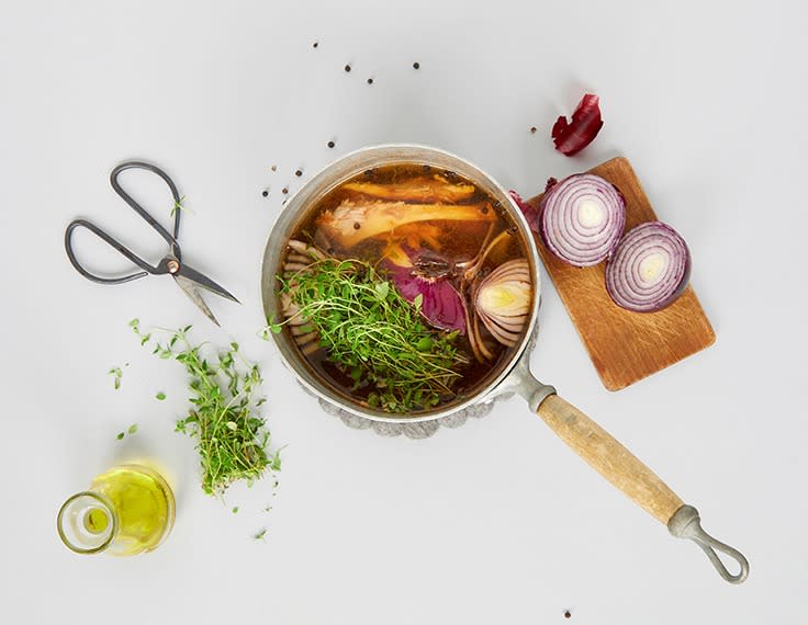 Hjemmelaget kyllingkraft i en liten kjele med ferske urter, og en delt rødløk ved siden av. En glassflaske med olje og en saks på siden.
