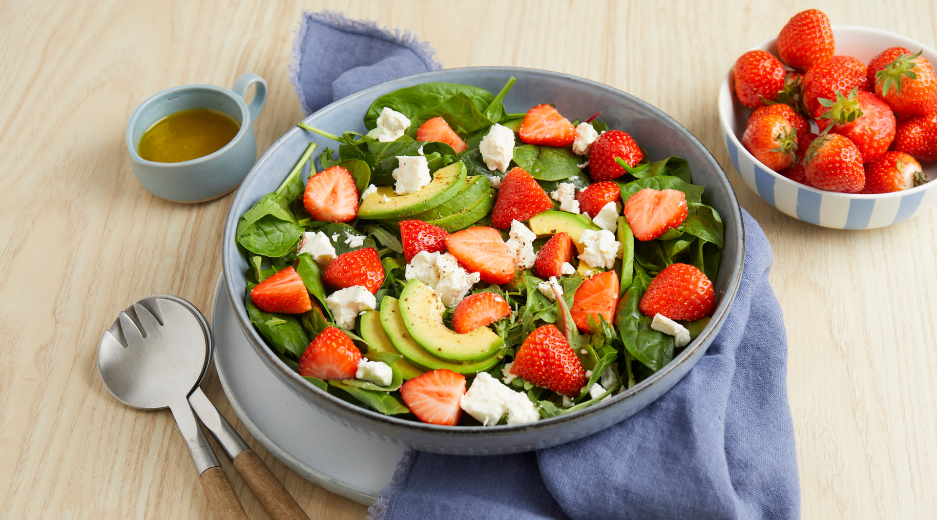 Jordbærsalat med fetaost og sitronvinaigrette servert i grå lav salatbolle med friske jordbær og avokado. Bollen står på lyst trebord med salatbestikk og en skål friske jordbær på siden, samt en liten mugge dressing bak.