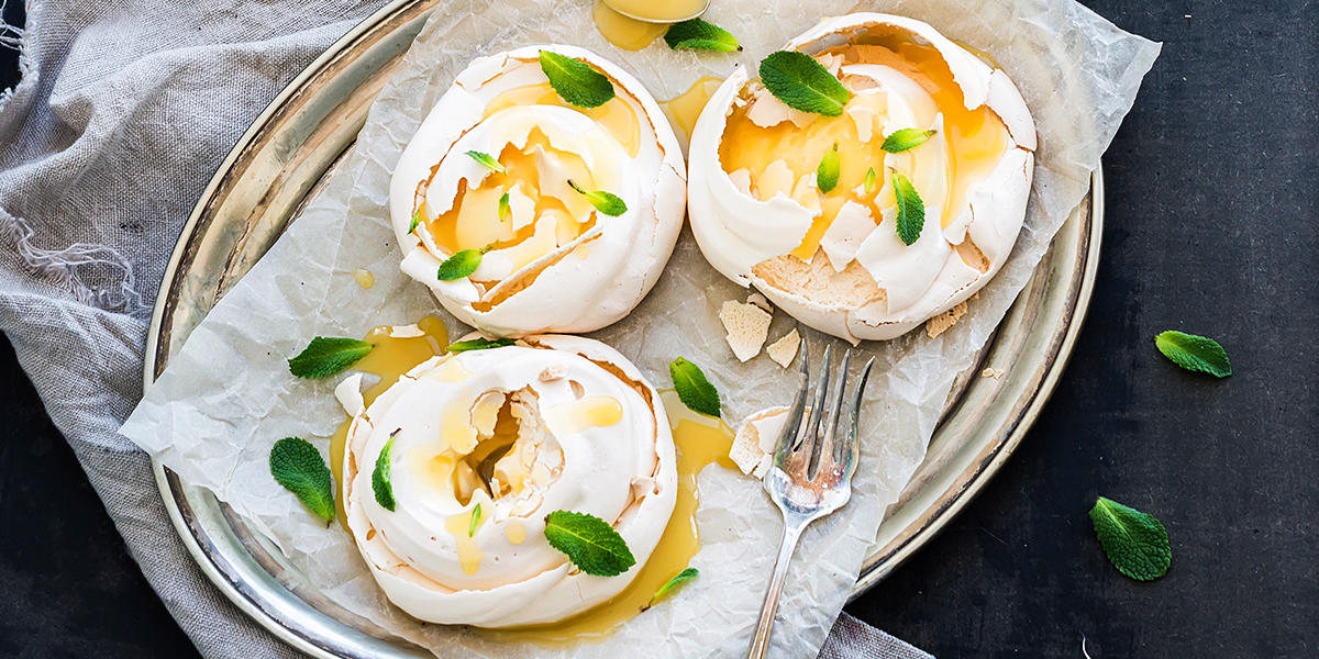 Mini pavlova med lemon curd og mynteblader, servert på sølvfat på mørk bakgrunn, med grå linserviett på siden.