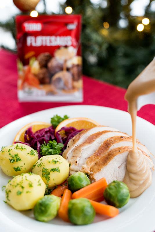 Tallerken med skiver av kalkun, poteter, gulrot og rosenkål på hvit tallerken. Fløtesaus blir helt fra et hvitt sausenebb over kalkunen. Pose med Toro fløtesaus i bakgrunnen. Juletre og rød duk.