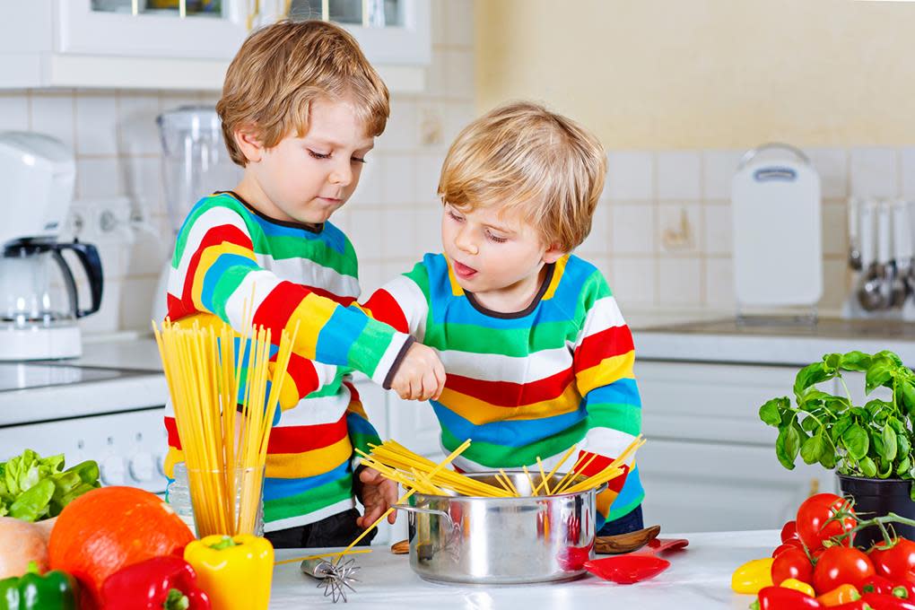 To små gutter med fargerike stripede gensere legger tørr spagetti i en kjele, på et lyst kjøkken fylt med grønnsaker.