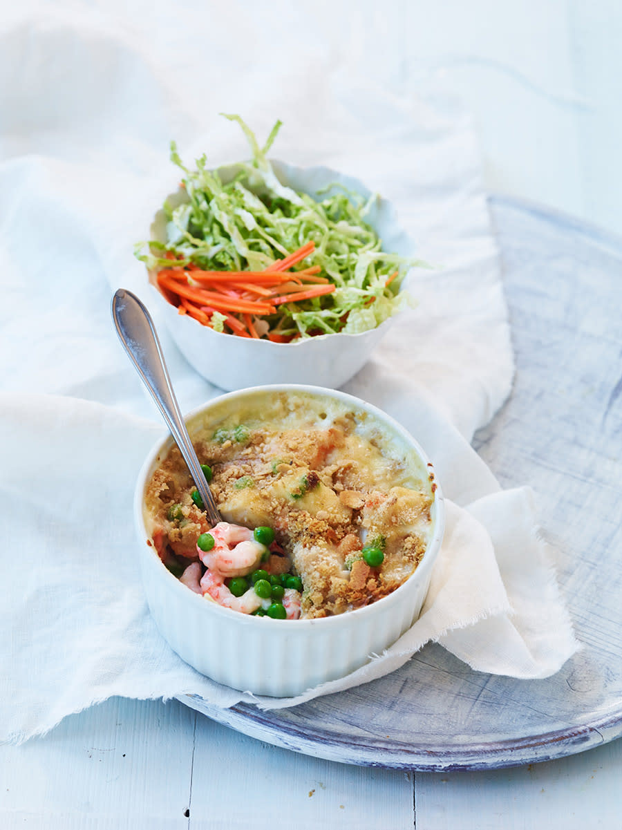 Enkel fiskegrateng med fiskepudding og reker laget i porsjonsform, og servert på hvit linserviett på lys tallerken. Hvit skål med råkost i bakgrunnen.