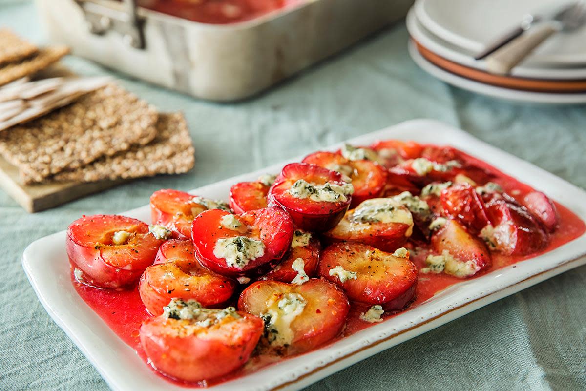 Ovnsbakte røde plommer med honning og blåmuggost servert på hvitt firkantet fat. Lys grønn linduk i bakgrunnen.