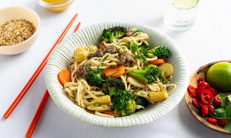 Wok med nudler, strimlet storfekjøtt og grønnsaker i en skål med røde spisepinner på venstre side, og en skål med chili, koriander og lime på høyre side