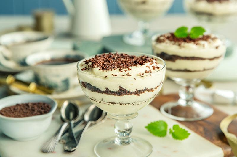 Dronning Maud dessert lagt lagvis i stettglass, og pyntet med et dryss revet sjokolade og noen blader sitronmelisse. Glassene står på hyggelig pyntet kaffebord.