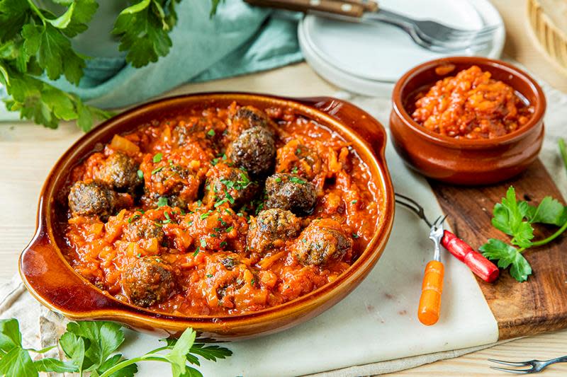 Hjemmelagede spanske kjøttboller til tapas i krydret tomatsaus, servert i brun ildfast form, på hvit marmorfjøl. Fersk bladpersille på siden.