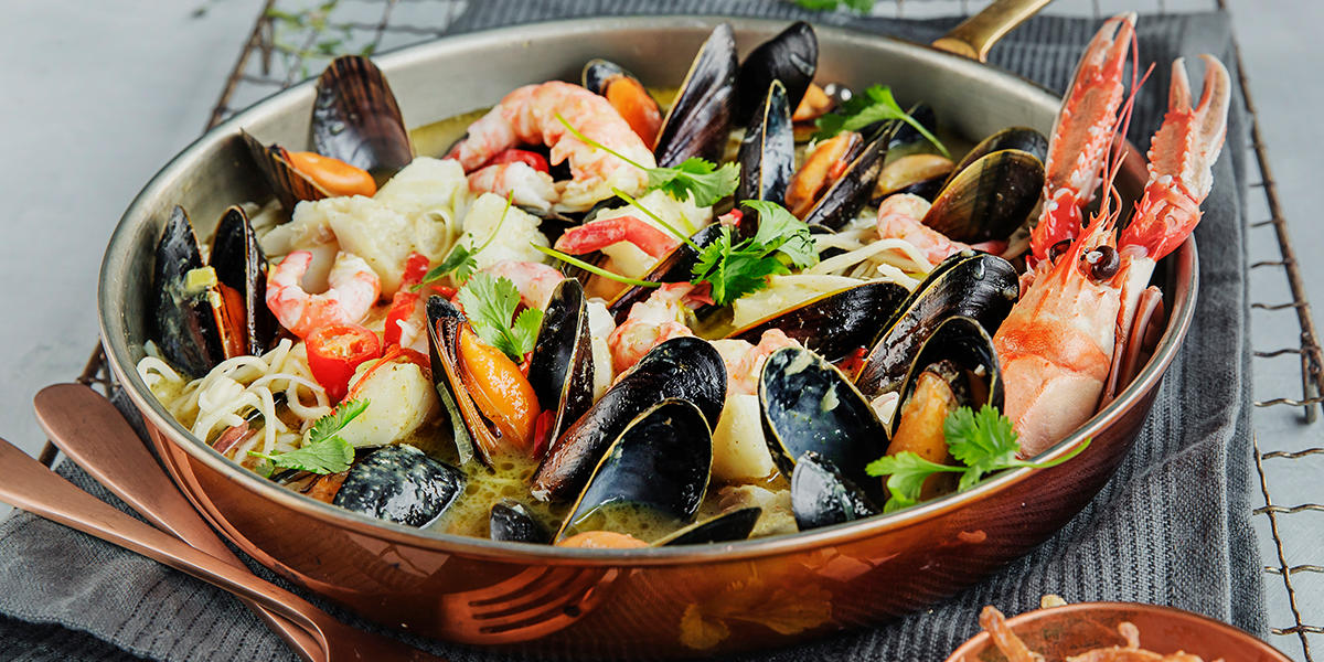 Asiatisk fiskegryte med sjøkreps, blåskjell, reker, nudler, chili og fersk koriander servert i kobbergryte på grått kjøkkenhåndkle.