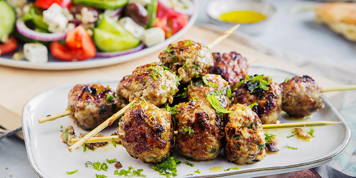 Fire spidd med små, stekte greske kjøttboller toppet med urter. Bak står en skål med gresk salat.