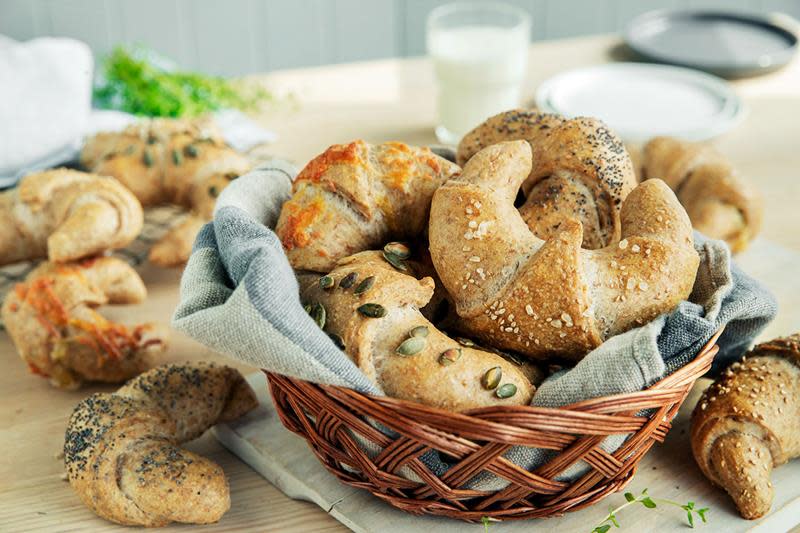 Grove horn med ost og skinke, toppet med forskjellige frø, ligger i en flettet kurv med blå linserviett, på et dekket kjøkkenbord.