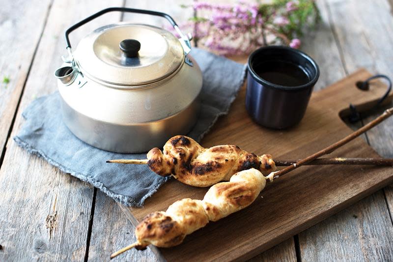 Turfavoritten pinnebrød på pinne, servert på trefjøl, ved siden av rustikk kaffekanne og kaffekopp. Skogsblomster i bakgrunnen.