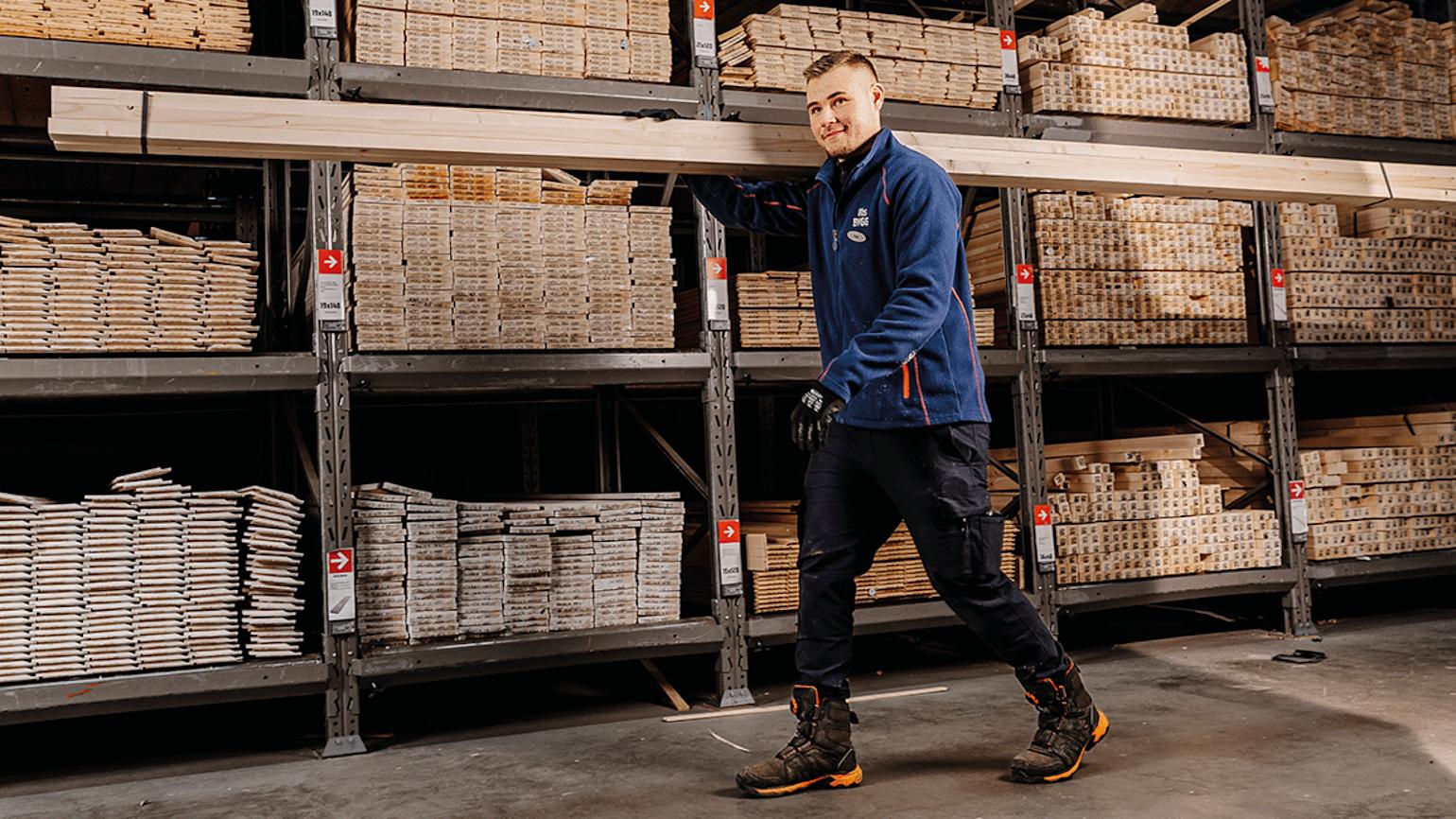 Mann smiler og går forbi planker i Obs Bygg butikk.