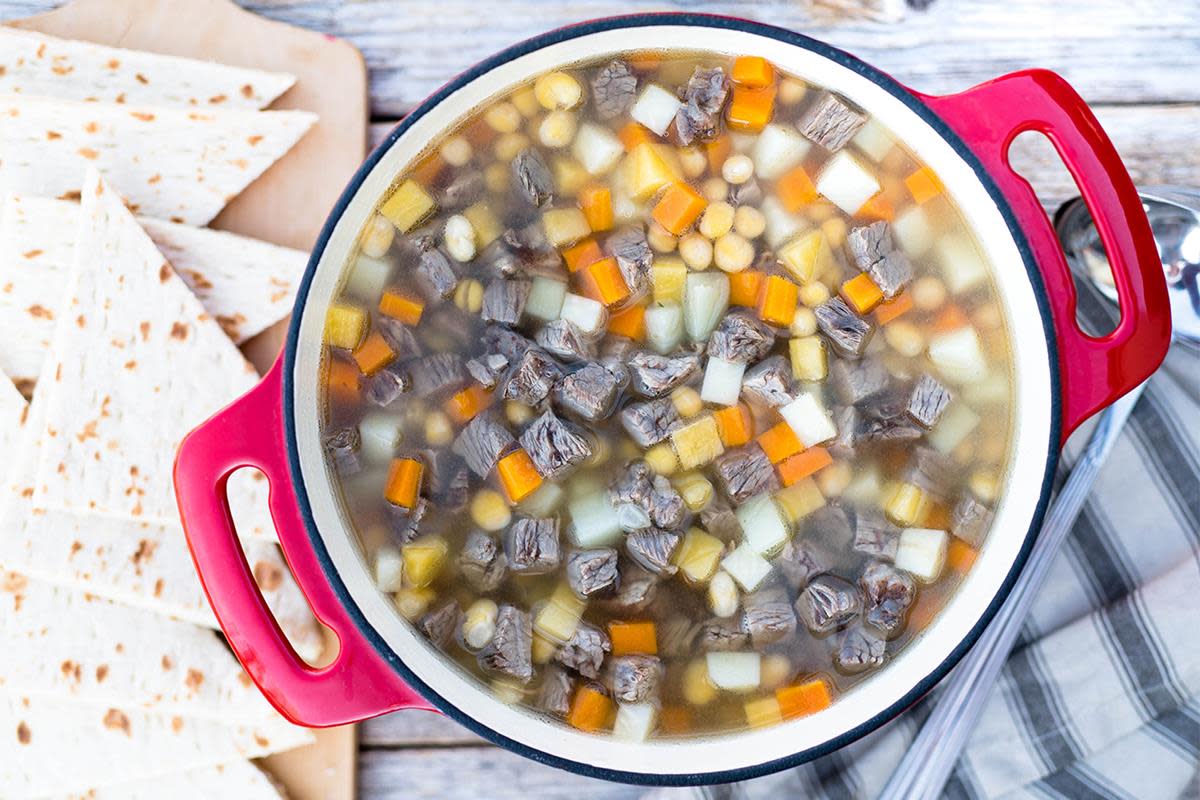 Kjøttsuppen Småmat med fargerike rotgrønnsaker og høyrygg i biter, koker i rød kjele. Servert på trebord med flatbrød ved siden av.