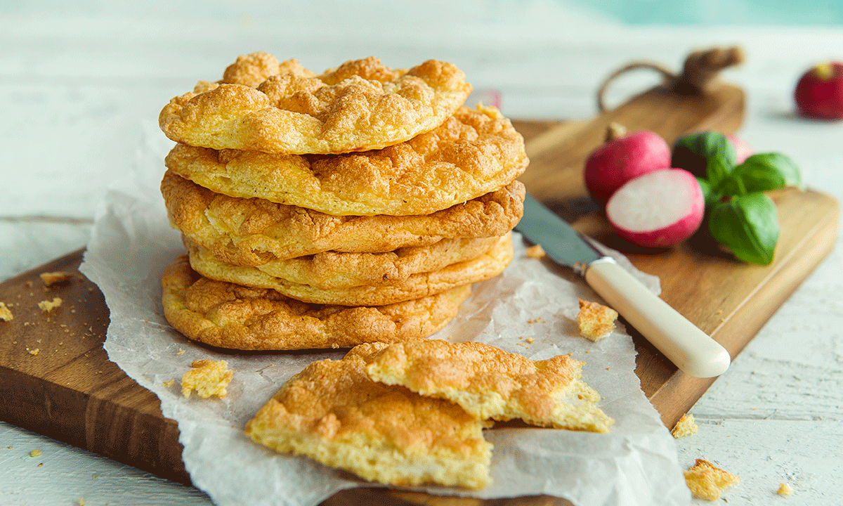 Cloud bread servert i stabel på kjøkkenfjøl i tre, med matpapir over. Små redikker, og en kvast frisk basilikum i bakgrunnen.