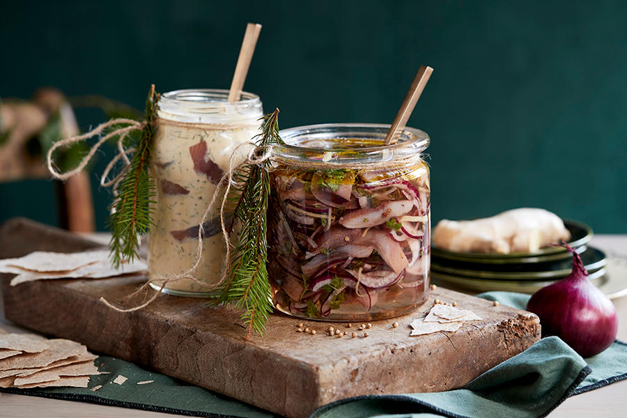 Hjemmelaget ingefærsild i glasskrukke på rustikk trefjøl med flatbrød, granbar, tørkede krydder, fersk rødløk og ingefær rundt. Smalere krukke med hjemmelaget sennepsild bak.