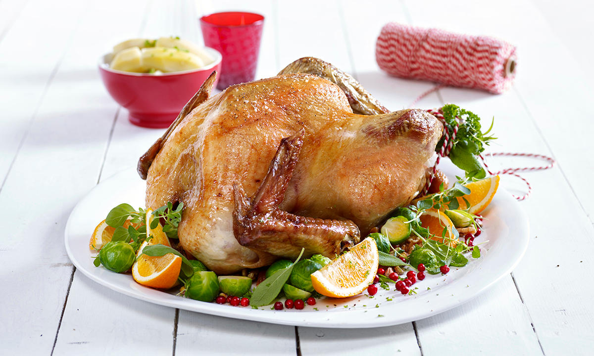 Supersaftig helstekt kalkun servert på hvitt fat med rosenkål, ferske urter og appelsinbåter. Fatet står på hvitt trebord, med rødt glass og rød hyssing i bakgrunnen.